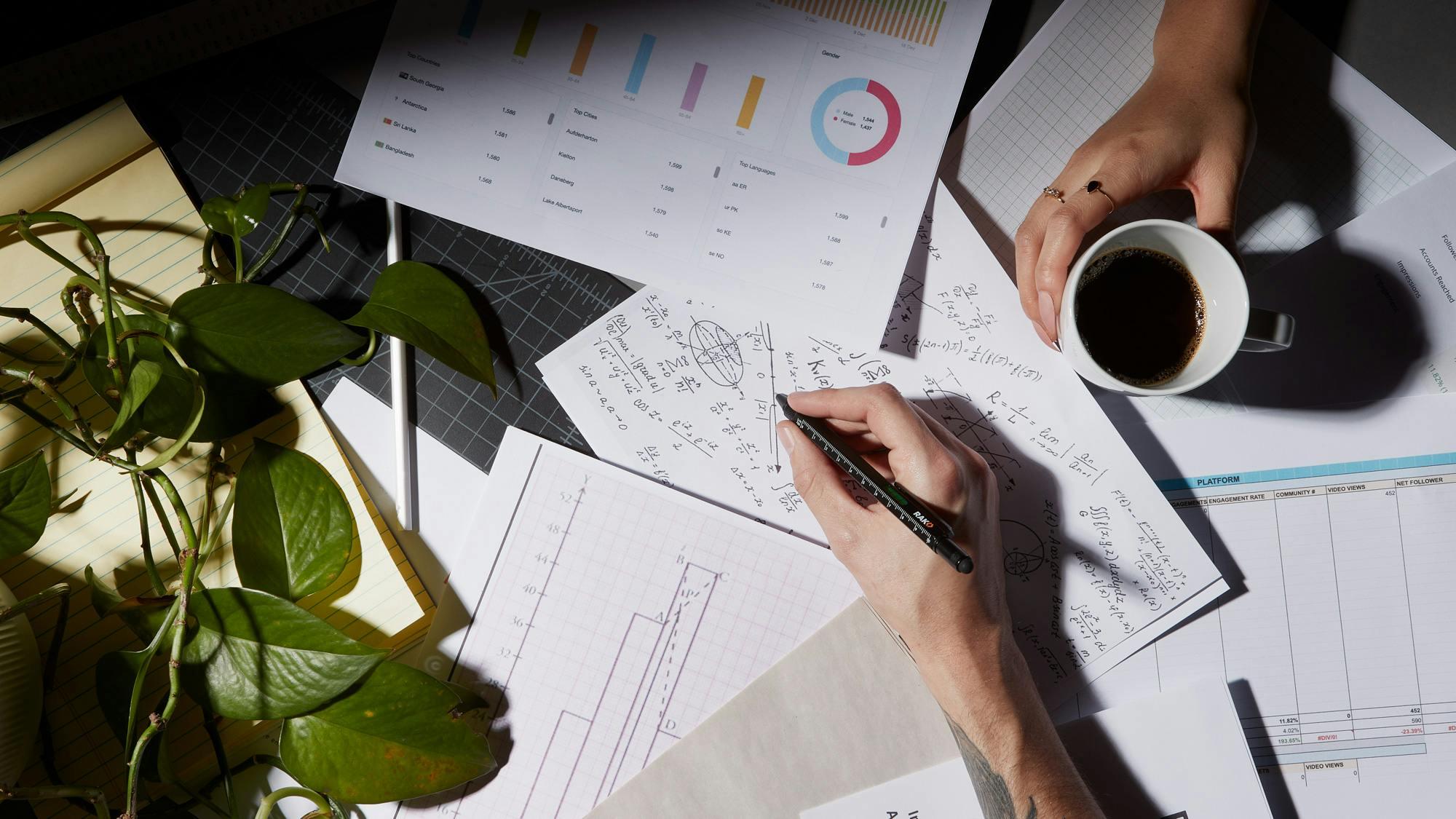 A hand holds a pencil, poised to write on a page filled with statistics and analytics, alongside a cup of coffee and a small plant. The page shows charts and graphs indicating data related to various metrics.