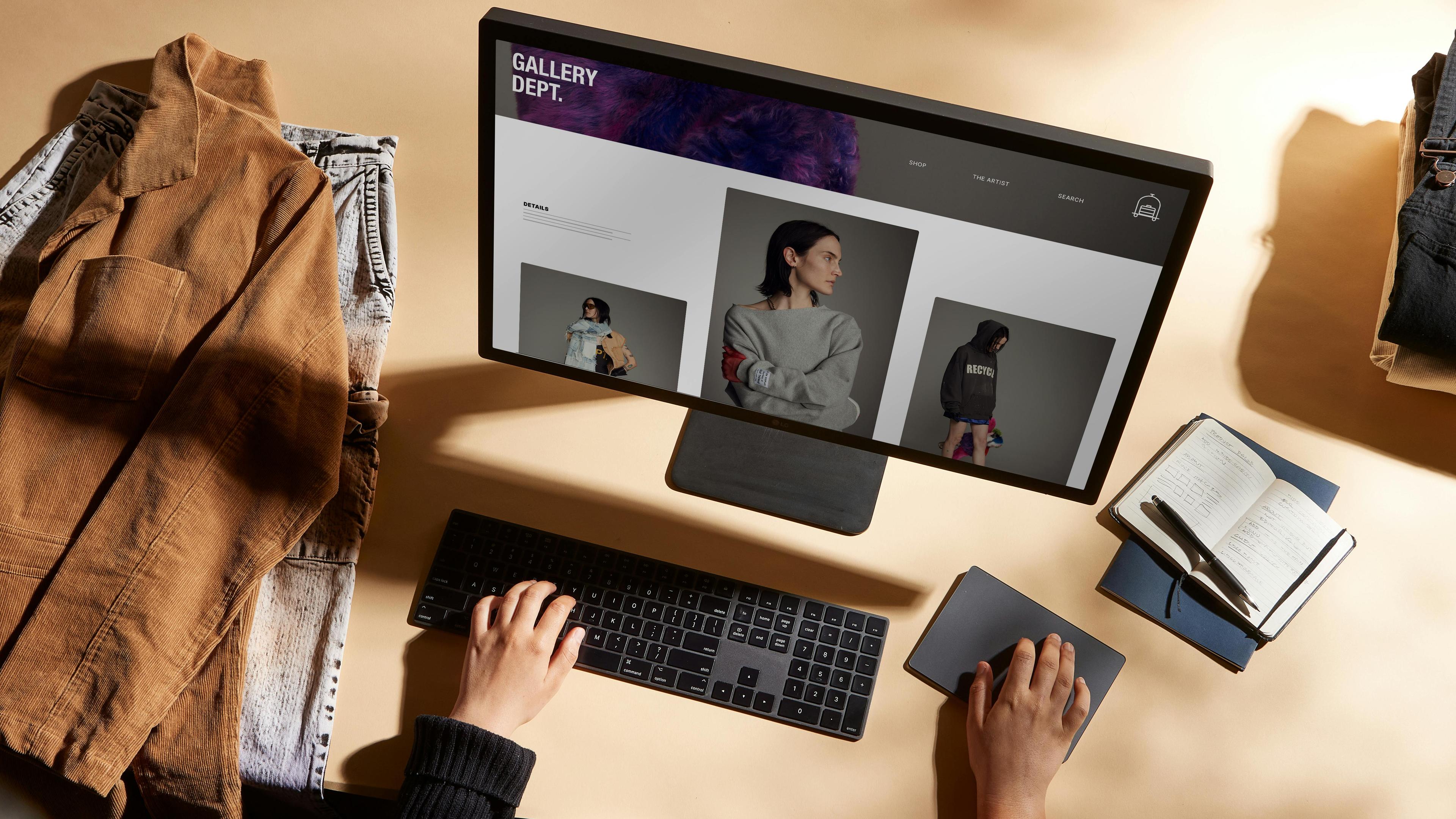 Ecomm Web development on computer monitor. Flat lay view of a laptop computer screen displaying a webpage, with a wireless keyboard and mouse, coffee cup, smartphone, and a notepad with a pen placed on a white desk. A pair of hands are visible on the sides of the keyboard, typing and clicking on the mouse