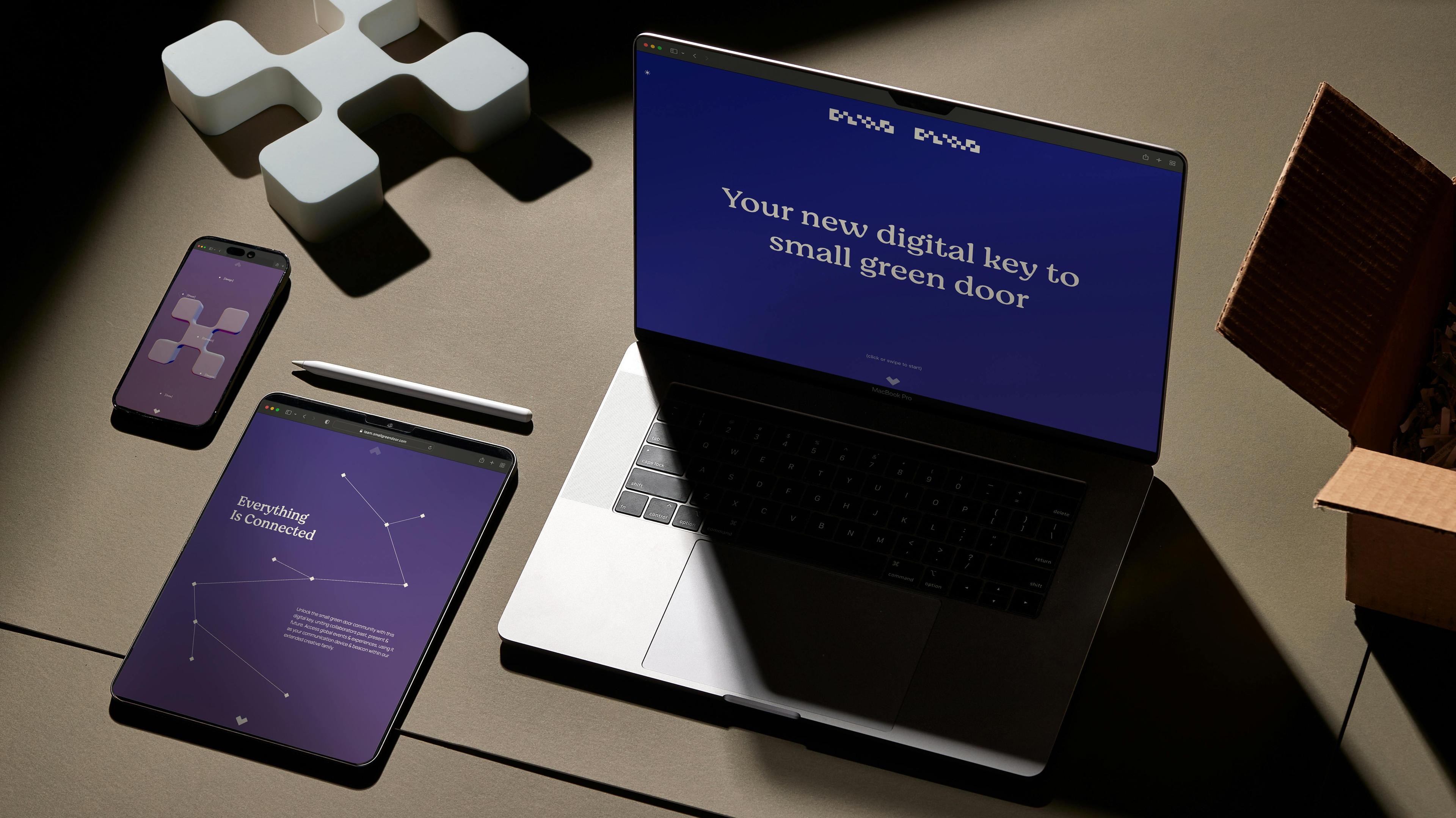 A flat lay of a laptop, iPad, iPhone, and other tech devices arranged on a wooden table. The laptop is open and displaying a design project, while the iPad and iPhone are showing mockups of a mobile app. This image represents the small green door digital and physical asset. 